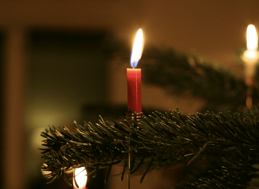 A small red candle burning on a Christmas tree branch, casting warm light in a dark room representing grieft and loss