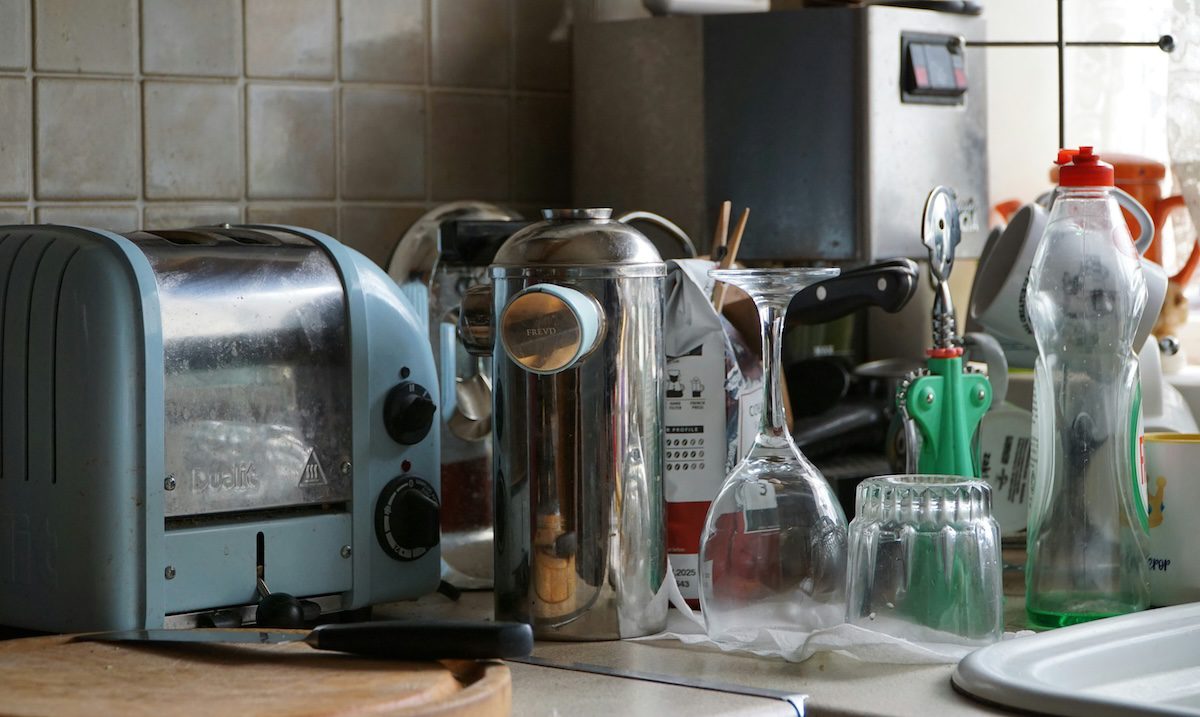 A cluttered kitchen countertop with scattered dishes, appliances, and glasses, creating a sense of overwhelm and household mess.