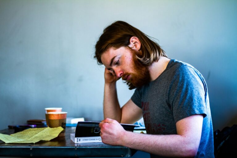 A person sitting at a table with coffee and papers, looking down while reading a book.